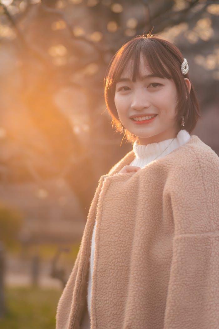 A young woman with a warm smile standing outdoors at sunset, wearing a stylish coat.