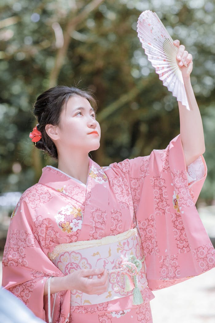 An elegant woman in a pink kimono holding a fan outdoors on a sunny day.